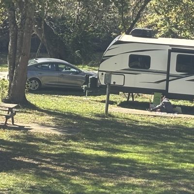 Two RVs parked under trees on grassy area, with a picnic table nearby.